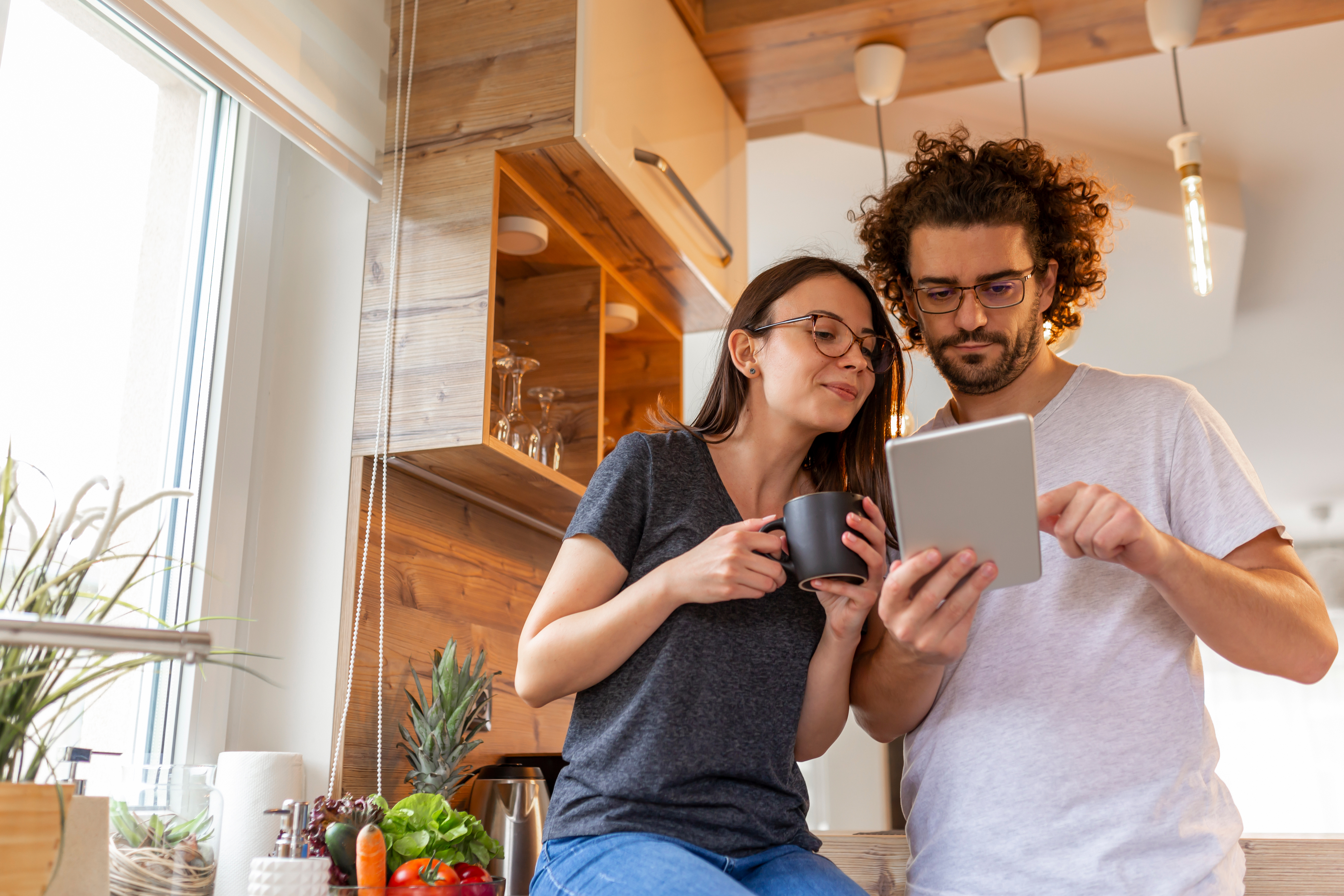 Eine Frau und ein Mann schauen auf ihr Tablet, sie stehen mit Kaffee in einer Küche.