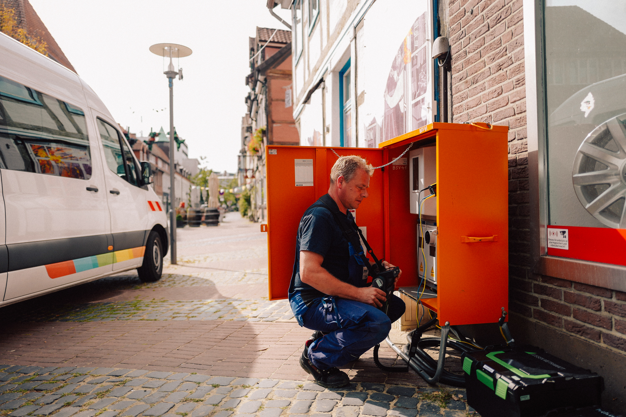Ein mycity Techniker sitzt an einem orangen Stromkasten und repariert ihn, neben ihm ein mycity Werkstattwagen