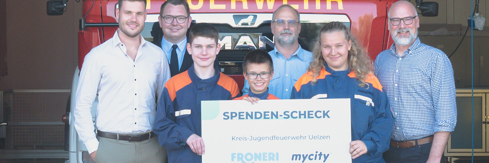 Vier Männer in schicker Kleidung stehen neben drei Kindern der freiwilligen Feuerwehr, die Kinder halten einen mycity & froneri Spenden Check über 1000 Euro in der Hand.