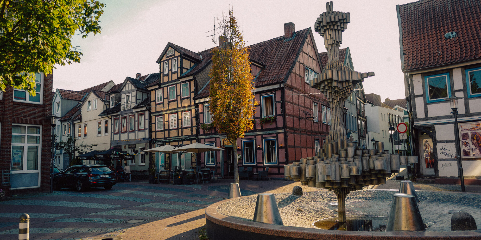 Eine bunte Häuser im Fachwerkstil stehen an einem Platz mit einem Baum darauf und einem großen künstlerischen Brunnen.