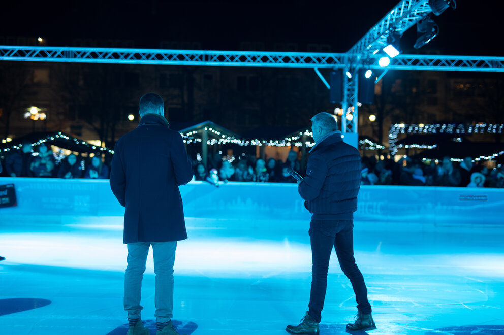 Eine Eisbahn mit mycity Schriftzug im Eis, zwei Männer stehen darauf und unterhalten sich mit einem Mikrofon, sie werden von hinten fotografiert.