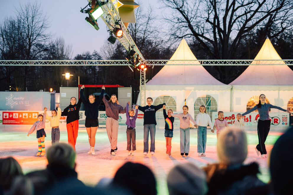Eine Gruppe Eiskunstläufer steht in einer Reihe und hält sich an den Händen und möchte sich vor Publikum verbeugen.