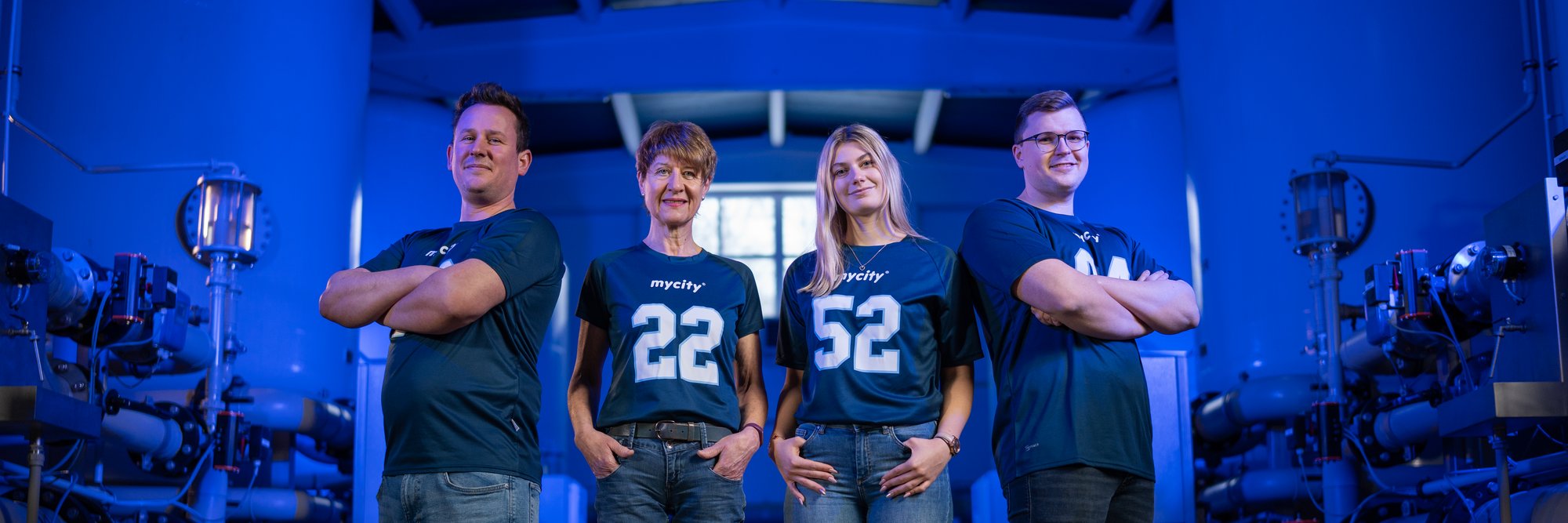 Zwei Frauen und zwei Männer stehen in blauen Shirts vor blauem Studiohintergrund, sie stellen ein Team dar.