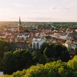 Luftaufnahme der Uelzener Skyline mit Häusern & Kirche zwischen vielen Bäumen, im Hintergrund ein hellblauer Horizont mit hellem Sonnenschein.