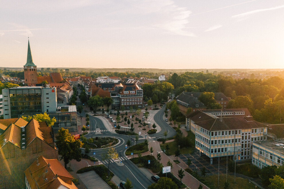 Luftaufnahme der Uelzener Skyline mit Häusern, Marktplatz & Kirche zwischen vielen Bäumen, im Hintergrund ein hellblauer Horizont mit hellem Sonnenschein.