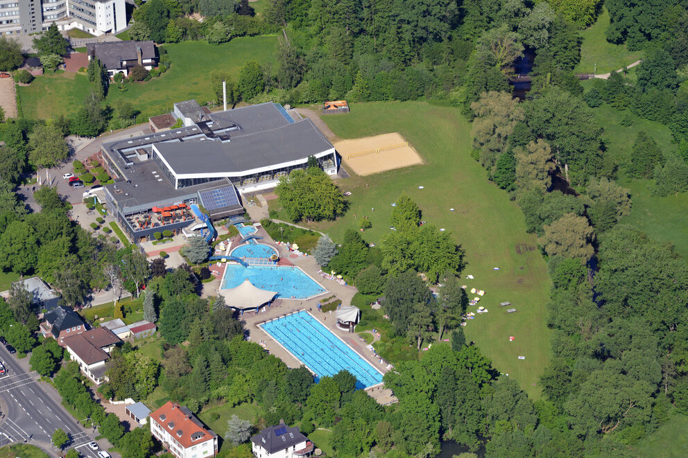 Luftaufnahme eines Schwimmbads mit mehreren Außenbecken in einem grünen Umfeld aus Bäumen und Wiese.