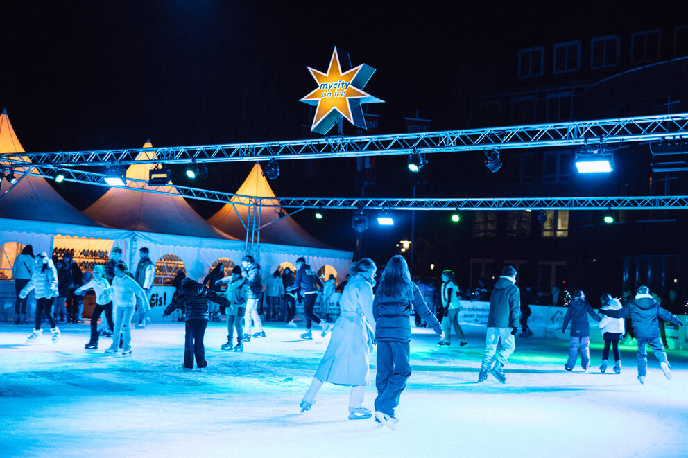 Einige Menschen fahren Schlittschuh auf einer Eisbahn. Über der Eisbahn ist ein Stern aufgehängt auf dem steht "mycity Eisbahn"