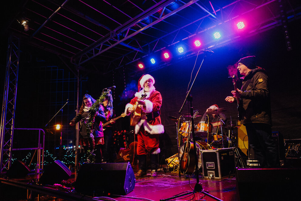 Ein Nikolaus steht mit einer Gitarre auf einer Bühne und eine Band steht auch dabei, sie singen ins Mikroi