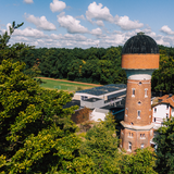Ein Wasserturm aus rotem Mauerwerk steht mittig zwischen einigen Gebäuden, ringsherum Wälder und ein kleines Fußballfeld