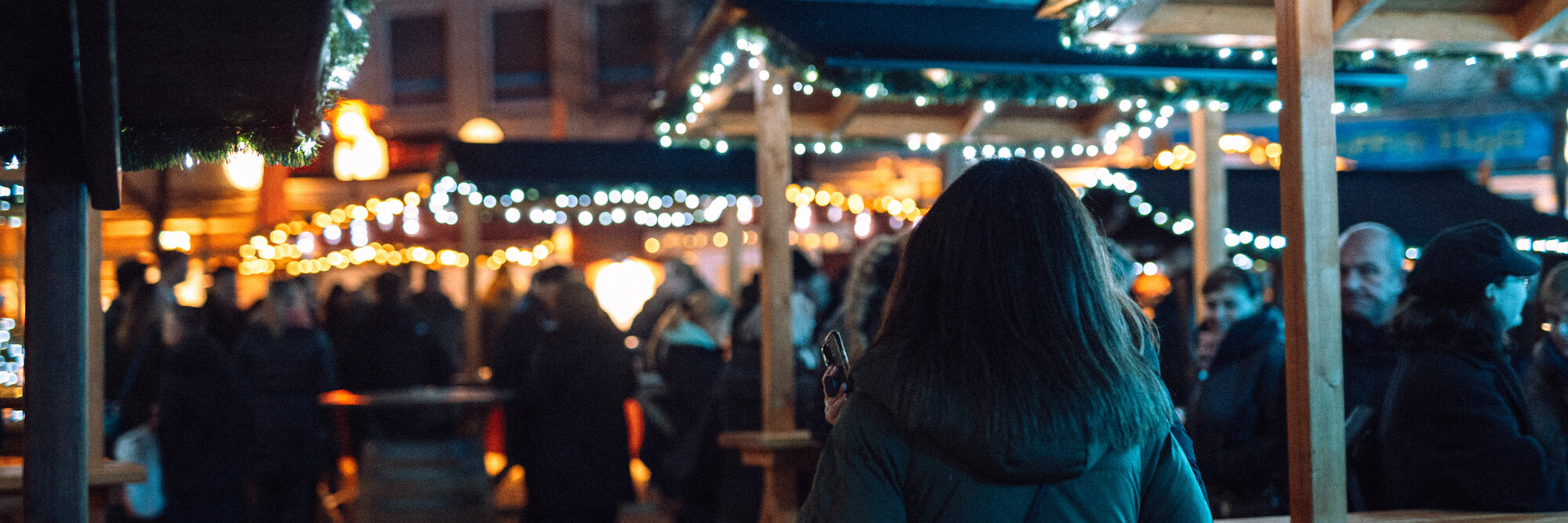 Ein Mädchen von hinten fotografiert steht vor einigen beleuchteten Ständen am Weihnachtsmarkt