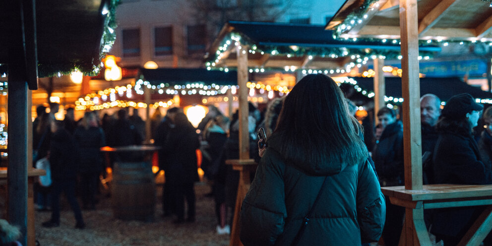 Ein Mädchen von hinten fotografiert steht vor einigen beleuchteten Ständen am Weihnachtsmarkt