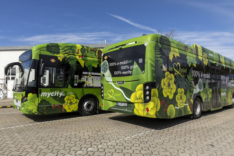 Zwei Busse von mycity mit grüner Beschriftung und Schriftzug 100% grün, ein Bus von vorne und ein Bus von hinten, seitliche Perspektive.