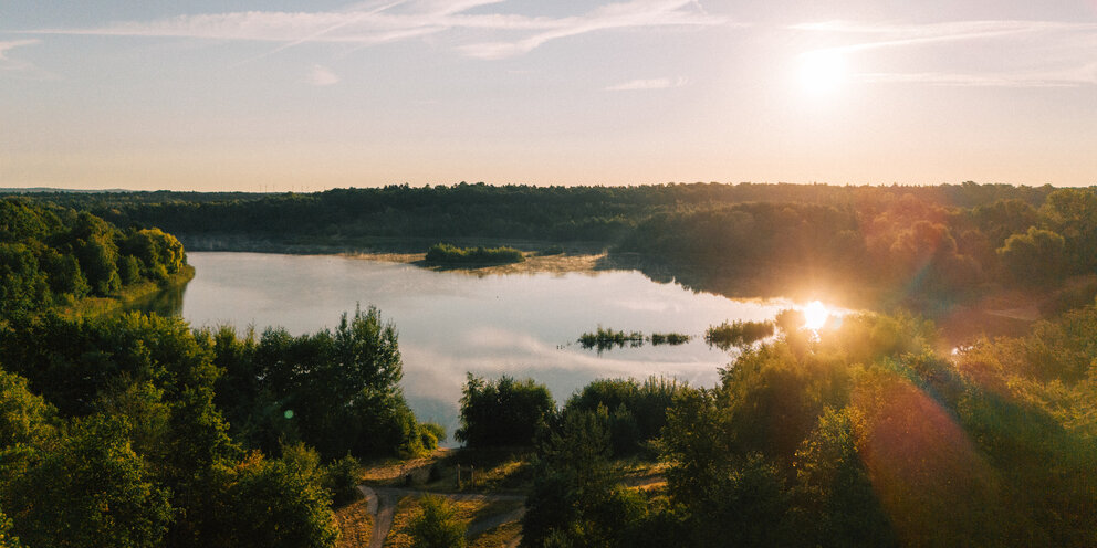 Ein großer See mit einem kleinen Weg und Wäldern herum, im Hintergrund ein hellblauer Horizont mit hellem Sonnenschein.