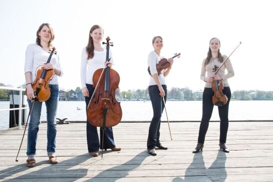 Vier Frauen in weißen Shirts und Jeans Hosen stehen auf einer Plattform vor einem See. Sie haben alle ein Sreichinstrument in der Hand.