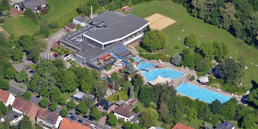 Luftaufnahme eines Schwimmbads mit mehreren Außenbecken in einem grünen Umfeld aus Bäumen und Wiese, vor dem Schwimmbad ist eine Straße und einige Häuser.