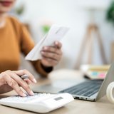 Eine Frau mit orangenem Shirt sitzt mit einem Taschenrechner & Block in der Hand an einem Laptop, dahinter eine Kaffeetasse.