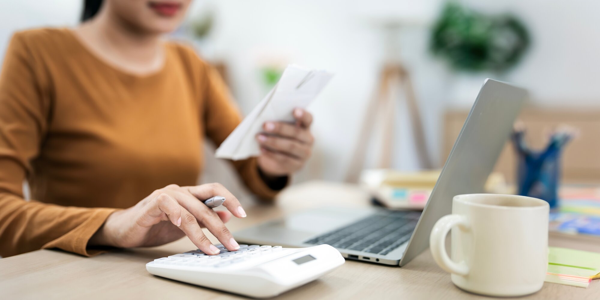 Eine Frau mit orangenem Shirt sitzt mit einem Taschenrechner & Block in der Hand an einem Laptop, dahinter eine Kaffeetasse.