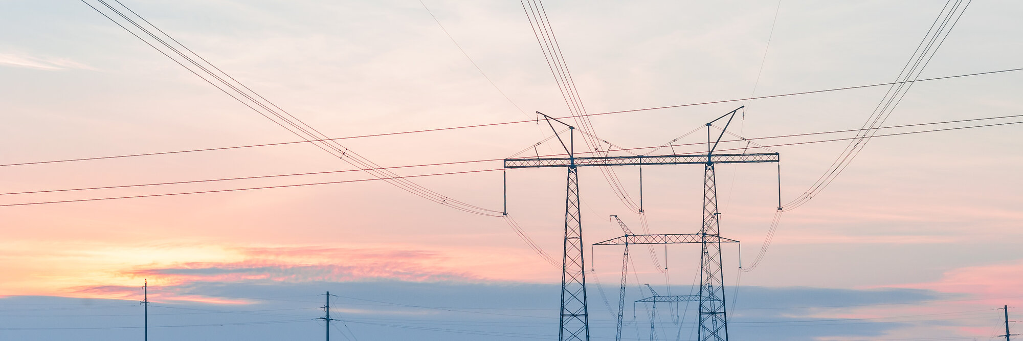 Eine Schneelandschaft mit Strommasten und leichtem orangenen Sonnenuntergang im Hintergrund.
