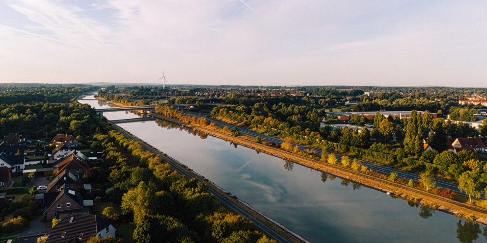 Ein großer Fluss mit Straßen und Bahnschienen daneben fließt zwischen vielen Bäumen und dazwischen sind einige Häuser.