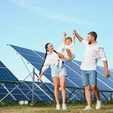 Ein Frau und ein Mann heben ihr Kind zwischen Ihnen an den Armen hoch und lassen es in der Luft fliegen. Im Hintergrund ein Solarpark.