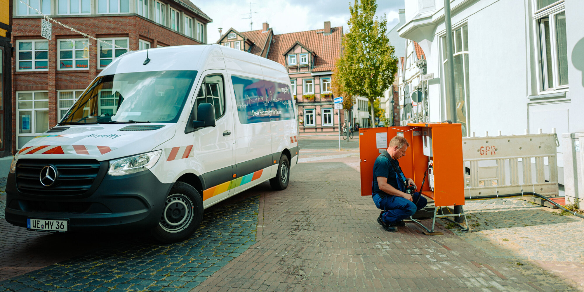 Ein mycity Techniker steht mit seinem Werkstattwagen an einem roten Stromkasten, er repariert diesen.