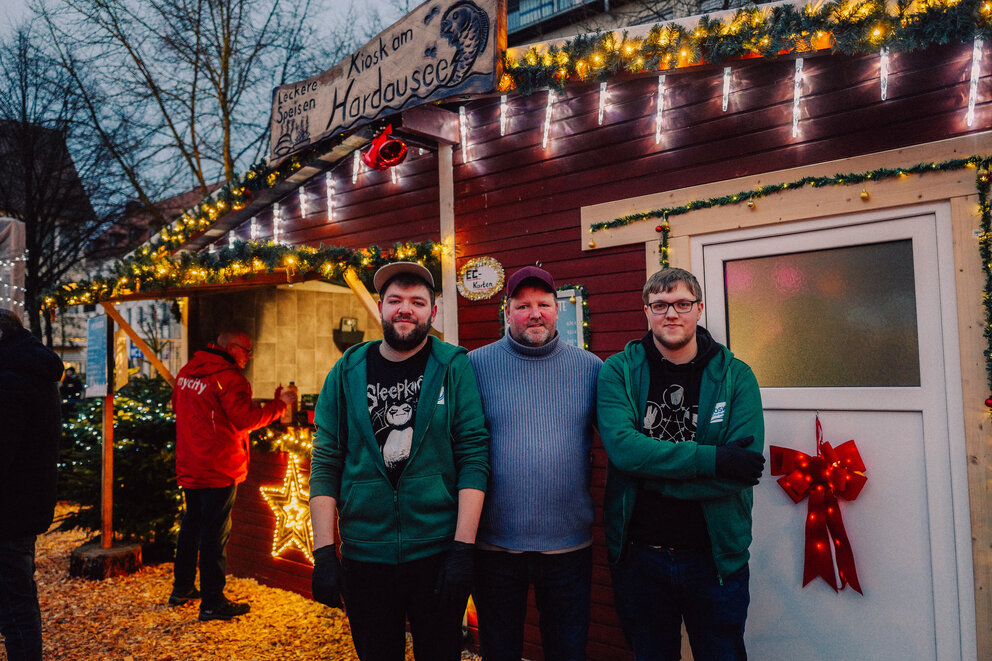 Drei Männer stehen vor einem Weihnachtsmarkt Stand mit der Aufschrift "Kiosk am Hardausee"