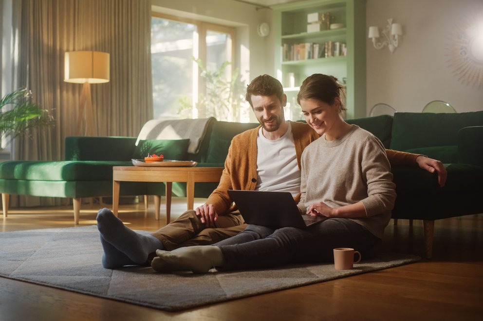 Eine Frau und ein Mann sitzen im Wohnzimmer vor ihrem Sofa auf dem Boden und schauen auf einen Laptop Bildschirm. Dabei lächeln sie.