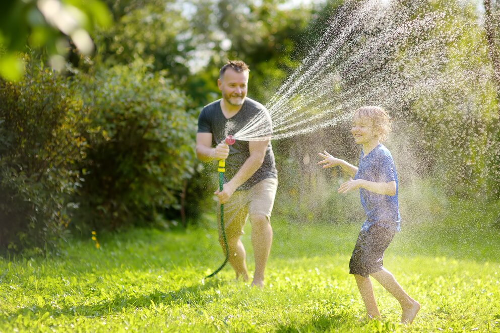 Ein erwachsener Mann und ein kleiner Junge spielen im Garten und spritzen sich mit einem Gartenschlauch und Wasser ab im Sommer.