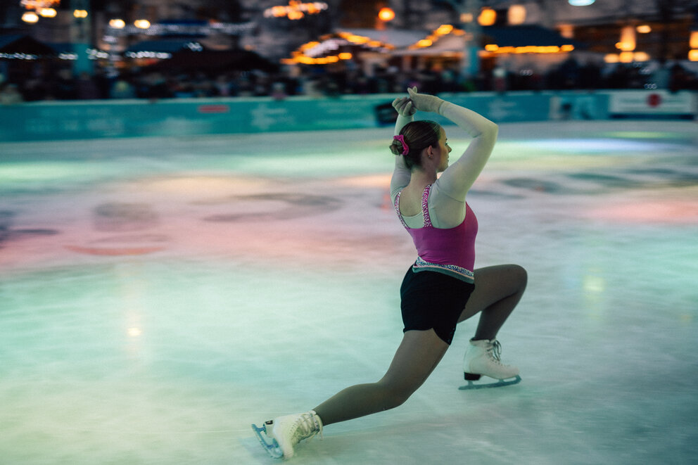 Eine Eisbahn mit einer Frau die eine Eiskunstlauf Vorführung präsentiert. Sie macht gerade eine Figur mit einem halb Spagat.