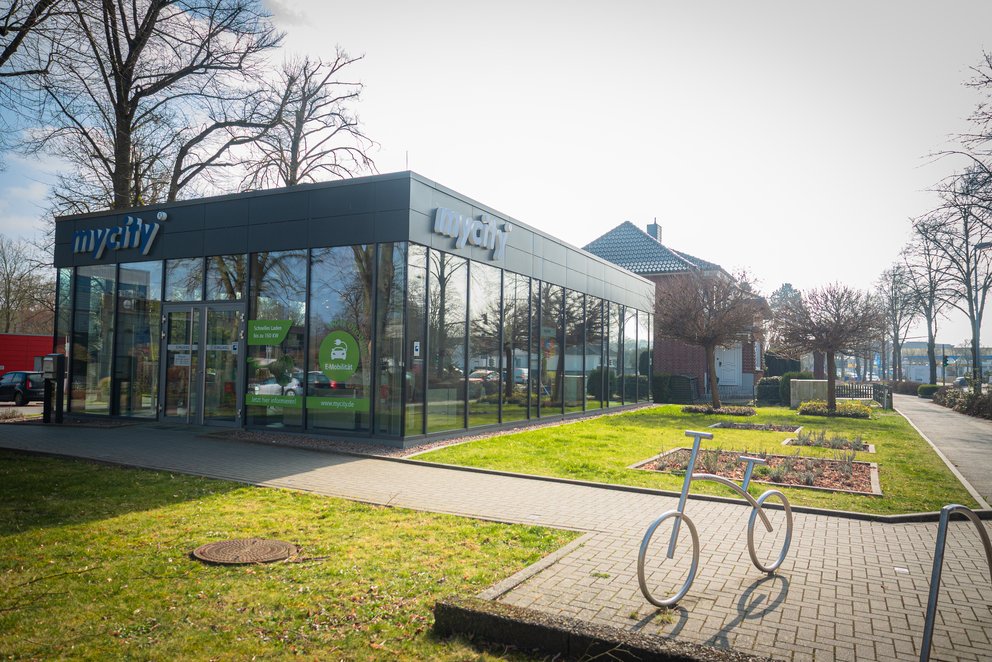 Ein komplett verglastes Gebäude, das mycity Service center mit einer Grünfläche davor und einem Fahrradständer, der wie ein Fahrrad aussieht.