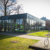 Ein komplett verglastes Gebäude, das mycity Service center mit einer Grünfläche davor und einem Fahrradständer, der wie ein Fahrrad aussieht.