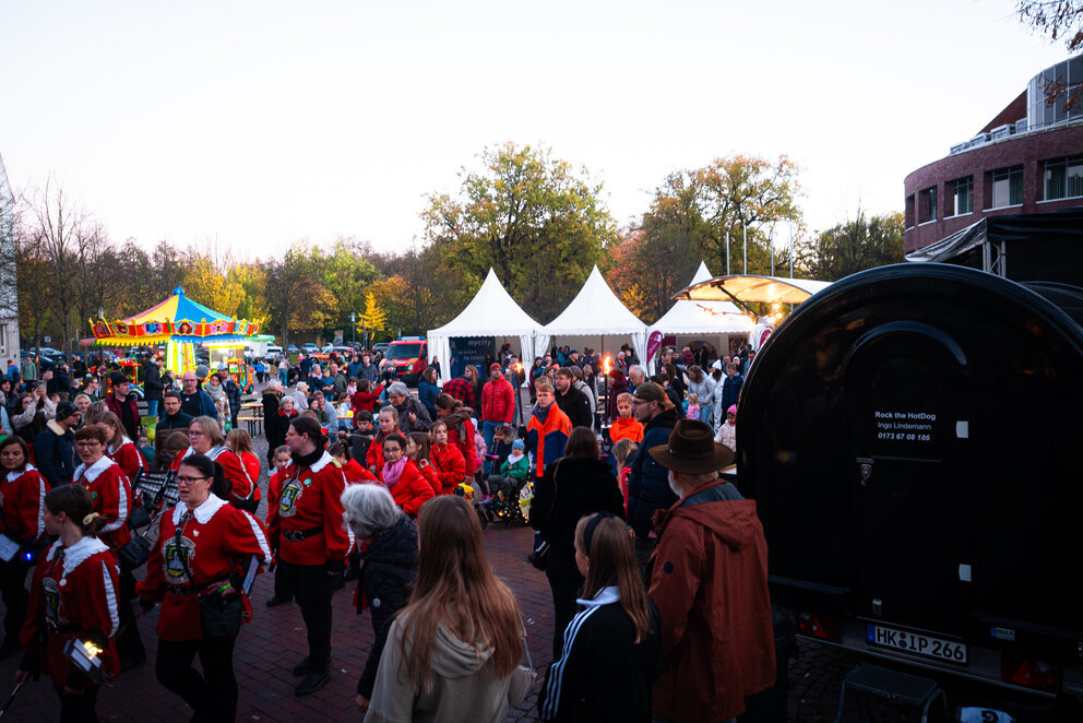 Ein großer Platz mit aufgebauten Partyzelten und Kinderkarusell auf dem viele Menschen sind, einige von Ihnen laufen in einer Reihe und tragen rote Mäntel.