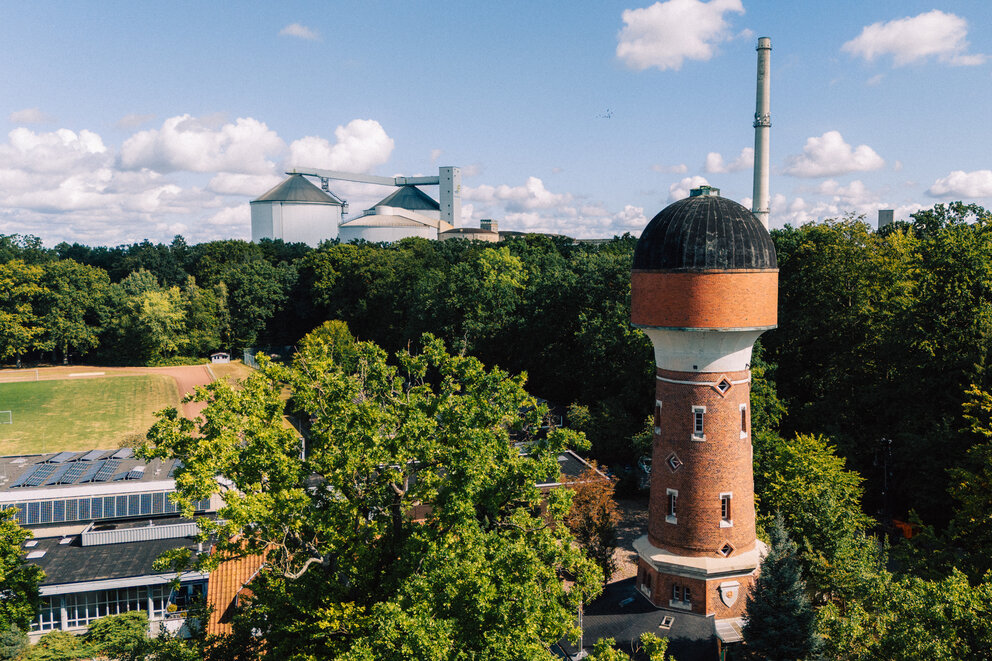 Ein Wasserturm aus rotem Mauerwerk steht links neben einigen Gebäuden, ringsherum Wälder und ein kleines Fußballfeld