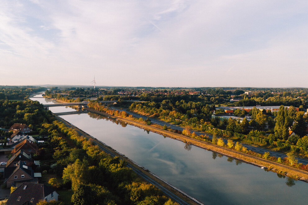 Ein großer Fluss mit Straßen und Bahnschienen daneben fließt zwischen vielen Bäumen und dazwischen sind einige Häuser.